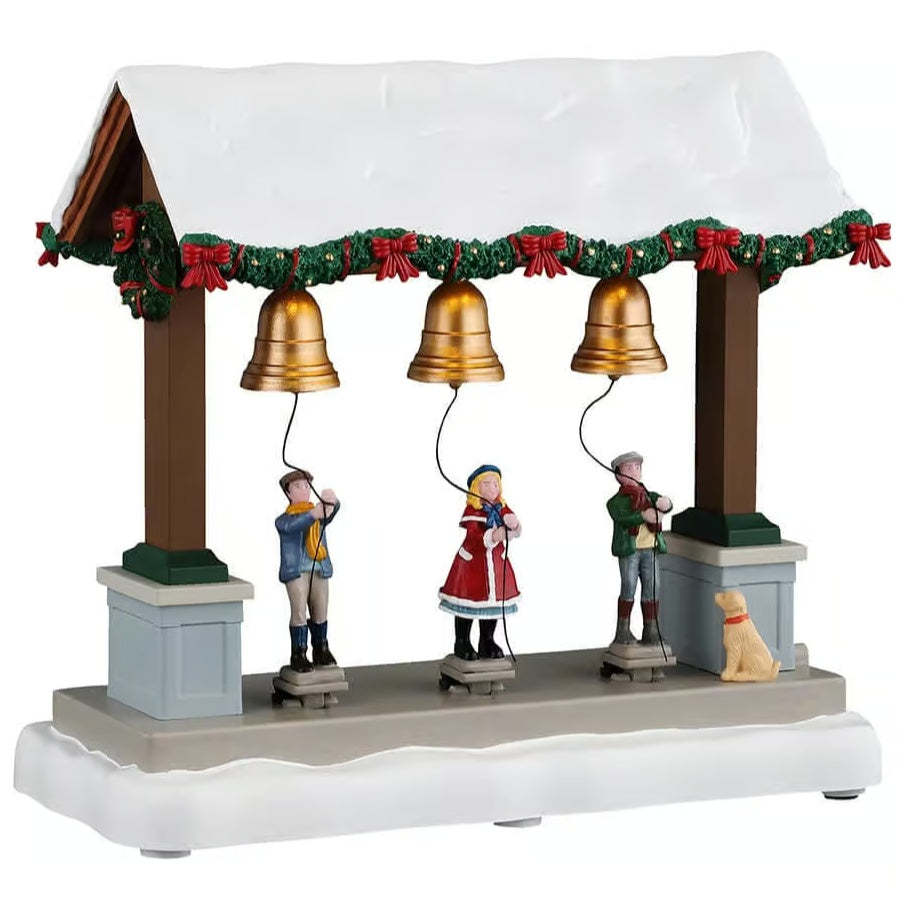 This image shows what this Lemax table accent looks like on a white background. It was designed to look like a wooden stall with three gold bells. The roof of the stall is covered in snow and garland decorated with red bows. There are three kids ringing the bells while a dog looks on.