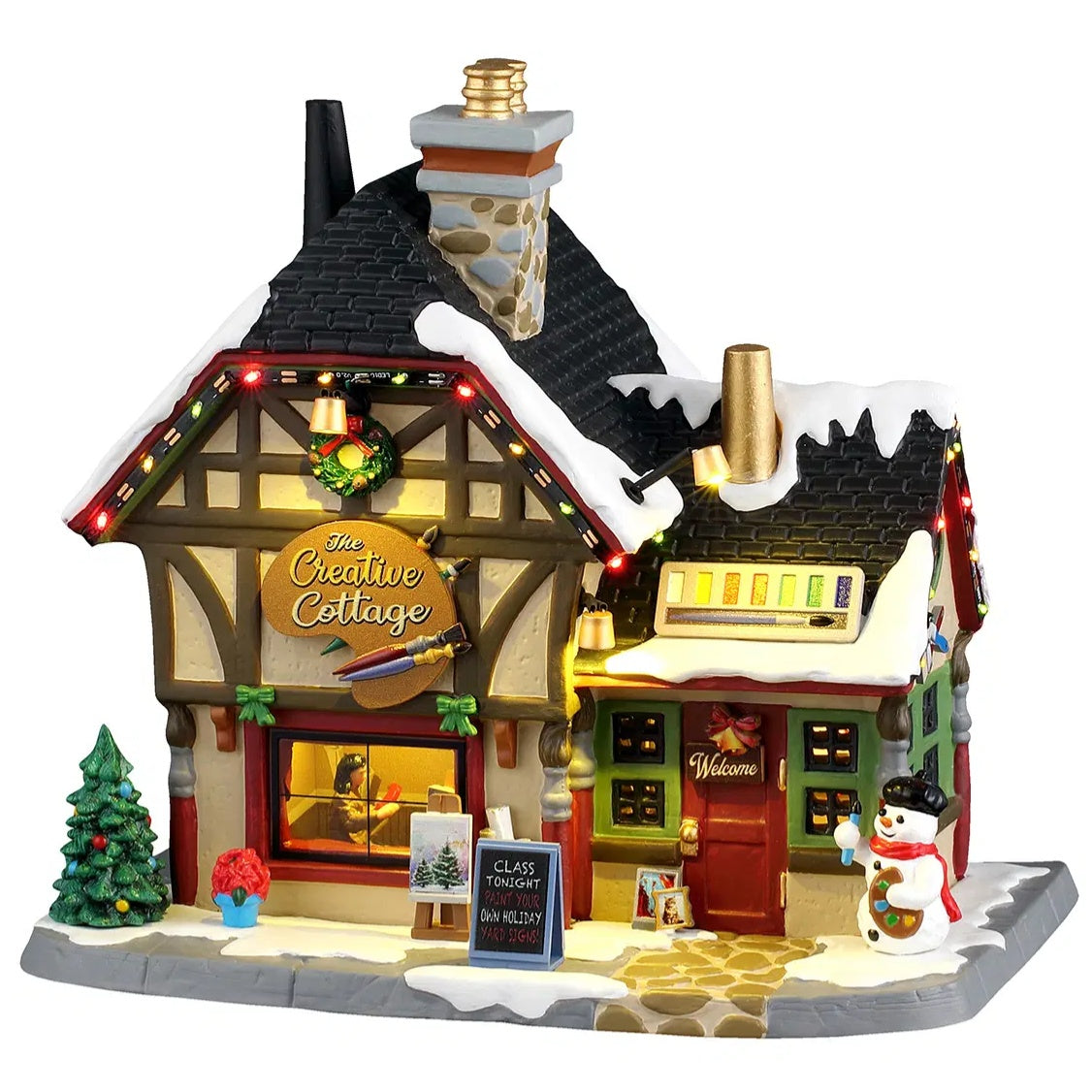 This image shows what this Lemax Caddington Village lighted building looks like on a white background. It was designed to look like a house turned into an art studio. The roof and the sidewalk has snow on it. Inside the window, you can see a woman painting. There's a decorated tree, a bouquet of flowers, an easel with a painting on it and a snowman on the sidewalk.
