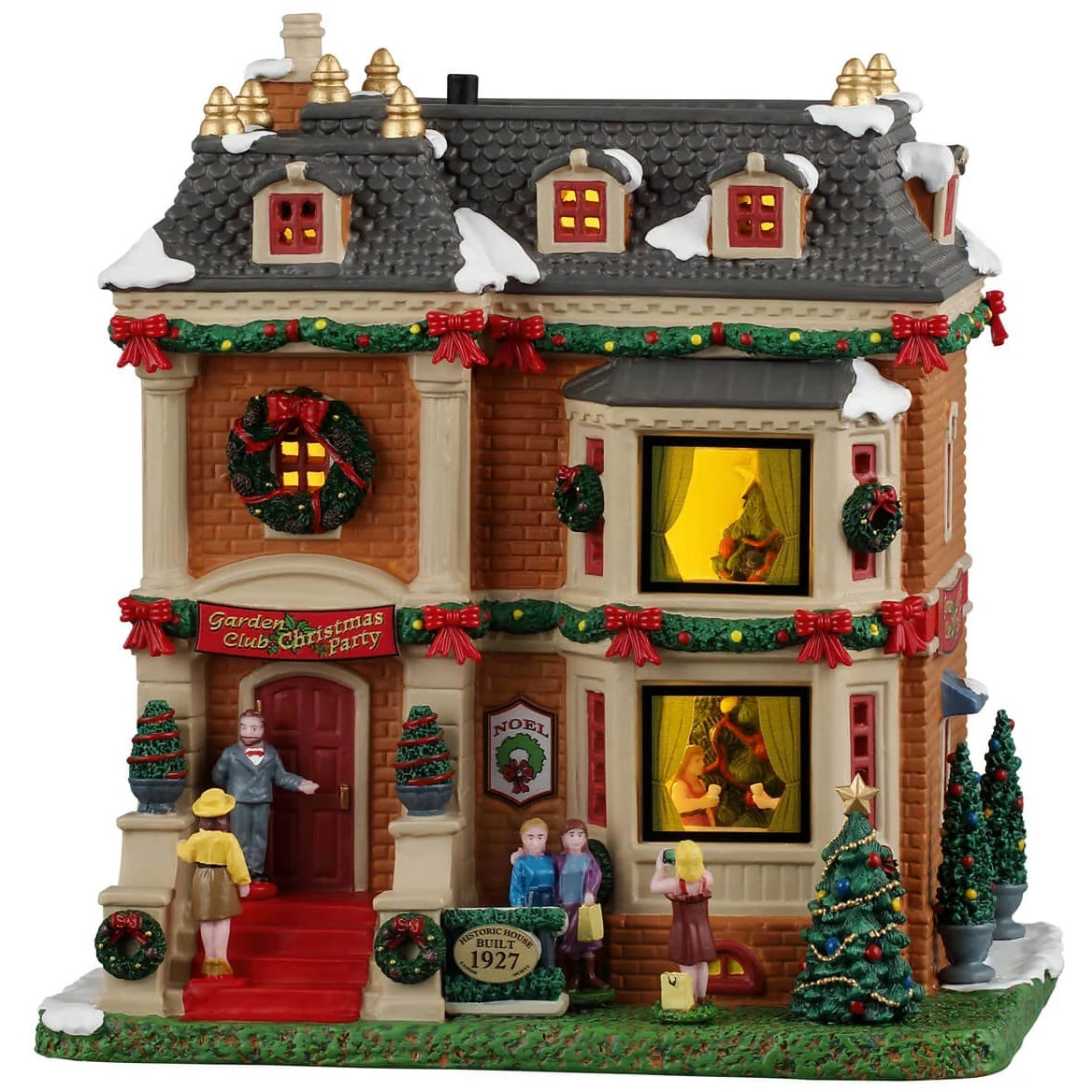 This image shows what this Lemax building looks like on a white background. It was designed to look like a tall Victorian-era style home. The rooflines are covered in green garlands with red bows. There's a large decorated wreath hanging above the front door. The potted plants and trees are also decorated with beads and ornaments. There are people taking pictures on the front lawn and a butler opening the front door for someone arriving to the party.