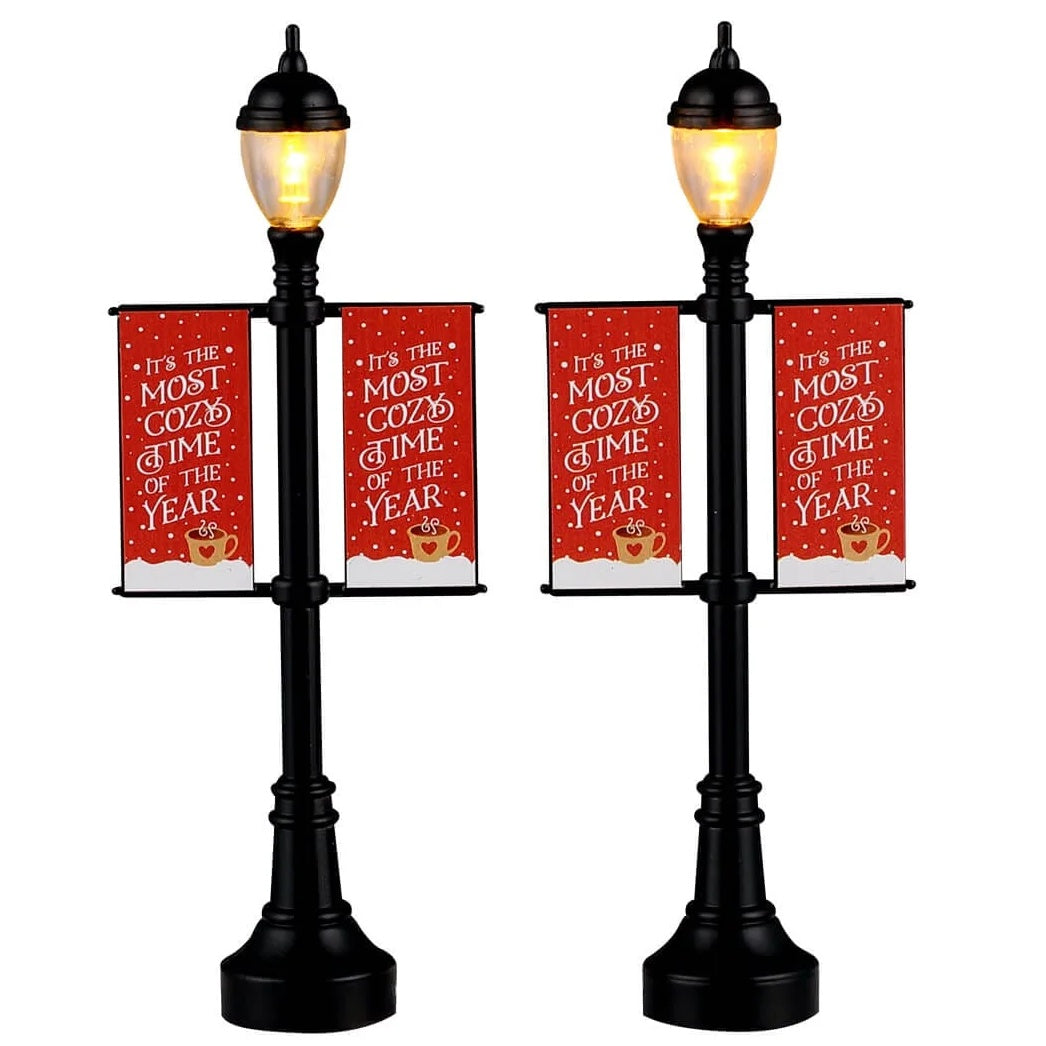 This image shows what these two Lemax Village accessories look like on a white background. They were designed to look like lamp posts decorated with banners for winter. The lamp posts are black. The banners re red and read "It's the Most Cozy Time of the Year."