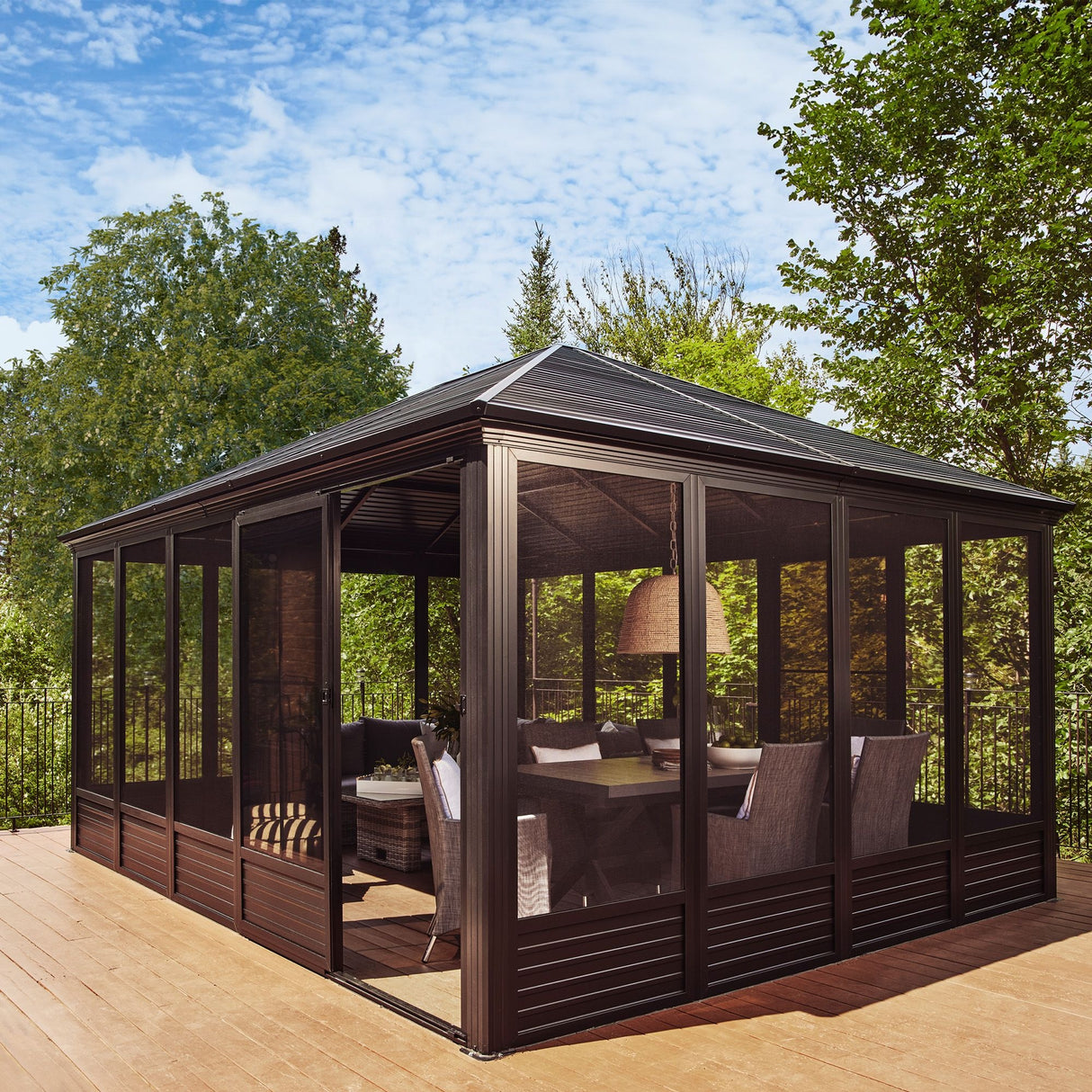 This image shows what his gazebo looks like on wooden deck on a very sunny day. Inside the gazebo, there are a few chairs, a dining table and big lamp.