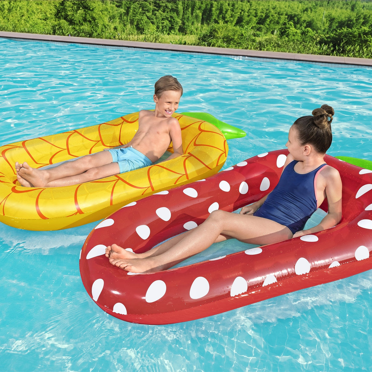 This is a lifestyle image showing two kids in floats in a pool. One boy is resting inside the pineapple float. One girl is resting inside the strawberry float.