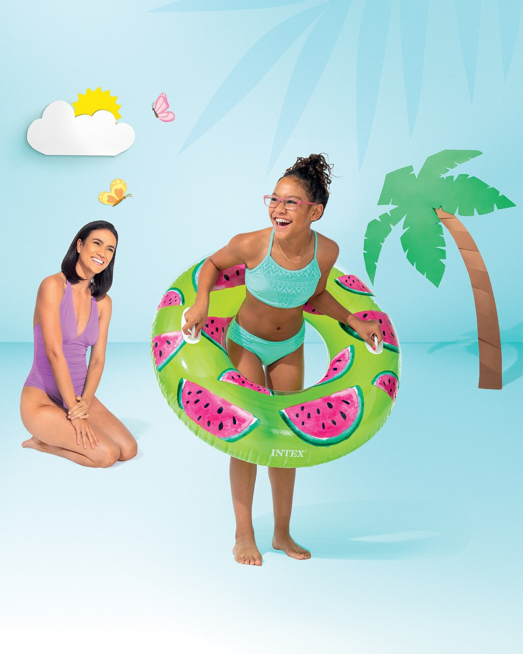 This is a lifestyle image showing a young girl standing inside the watermelon inner tube on a set that is designed to look like a beach made out of paper.