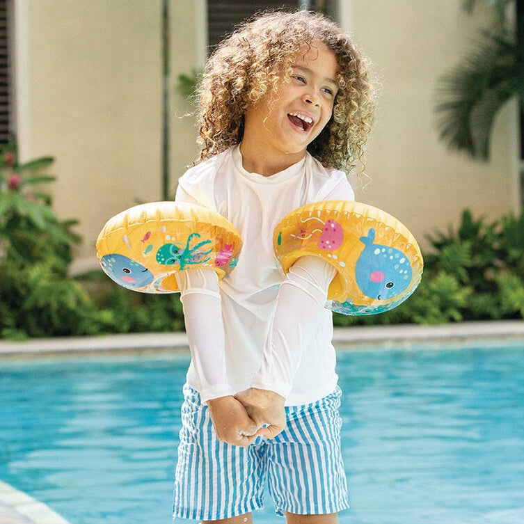 This is a lifestyle image showing a young child wearing both of these floaties on their arms on the side of a pool.