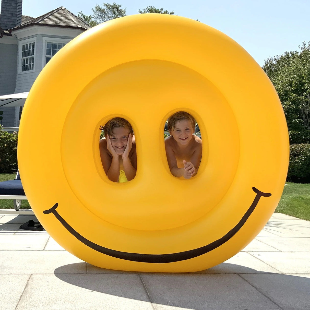 This is a lifestyle image showing the smiley face pool float standing upright on the sidewalk. There's a little boy in the left eye and a little boy in the right eye. They're looking at the camera and smiling.