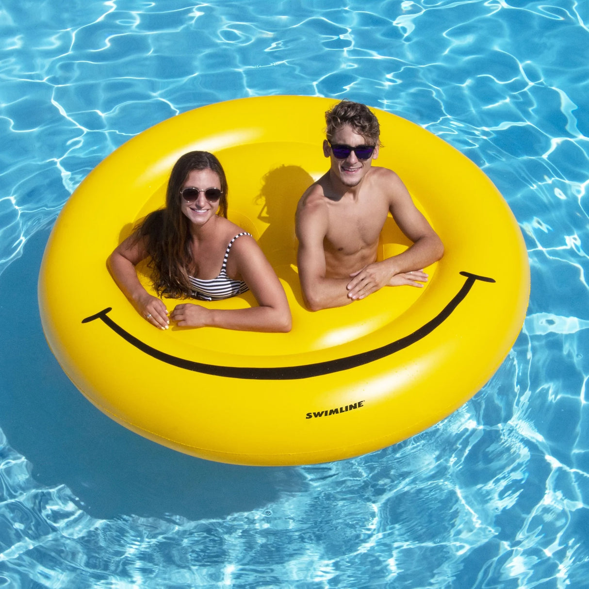 This is a lifestyle image showing the smiley face pool inflatable in the water. There's a girl wearing sunglasses in the left "eye" hole. There's a boy wearing sunglasses in the right "eye" hole.