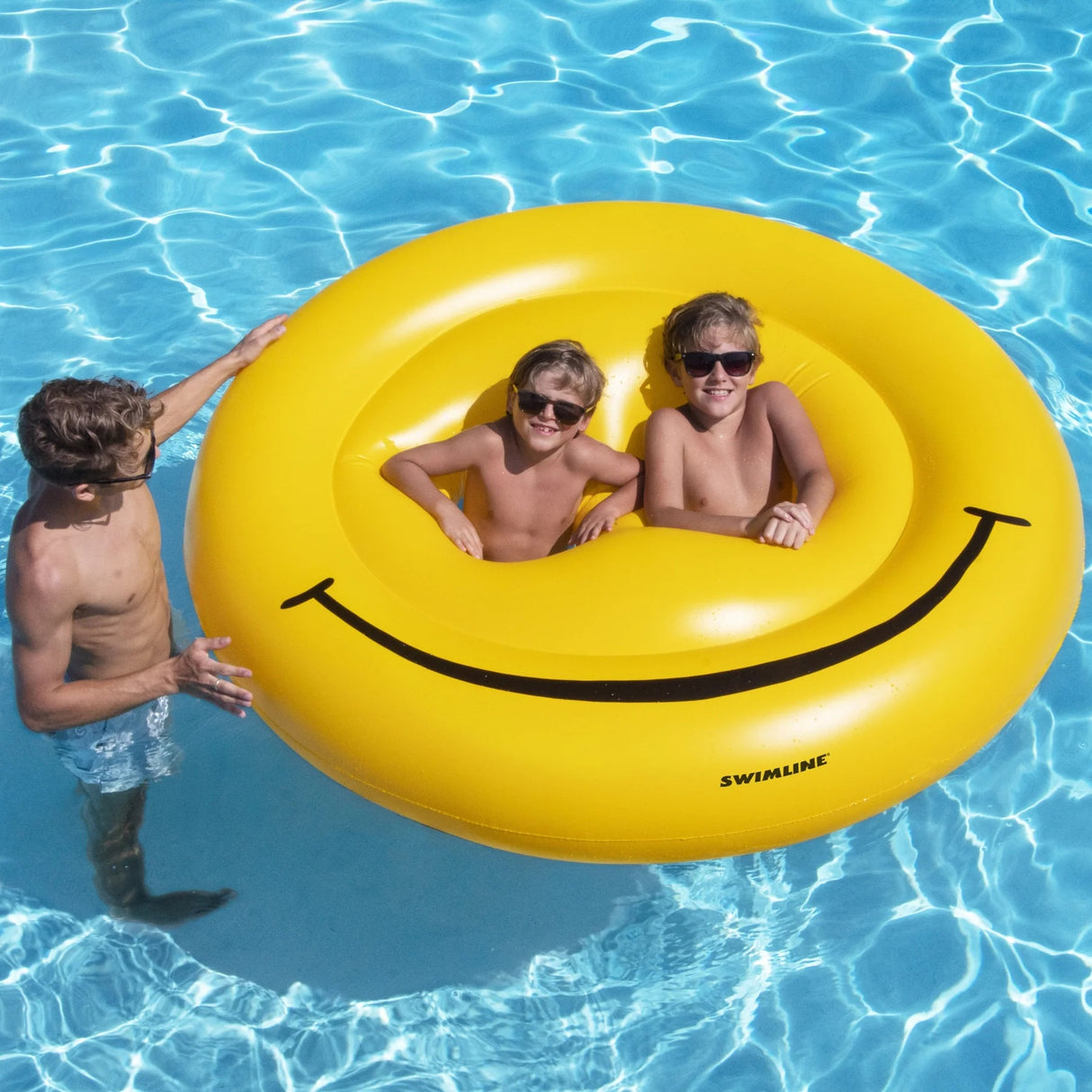 This is a lifestyle image showing two boys inside the "eye" holes of the smiley face inflatable. There's a dad holding the side of the float. The boys are wearing sunglasses and smiling at the camera.