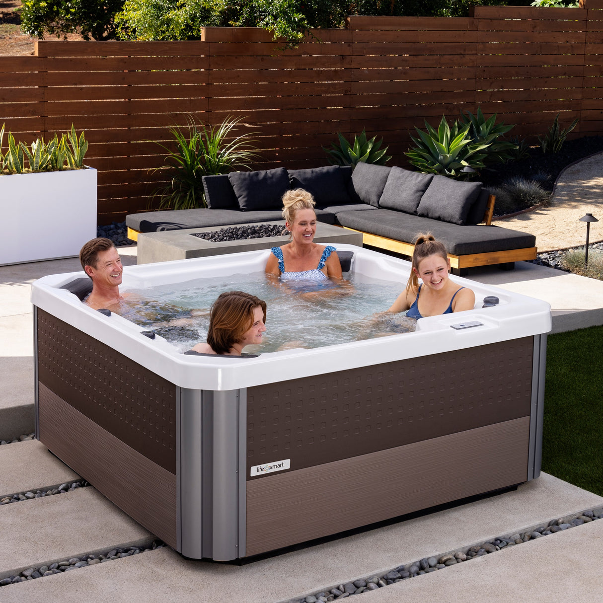 This is a lifestyle image showing four people in the hot tub on a patio next to a fire pit table and an outdoor couch.