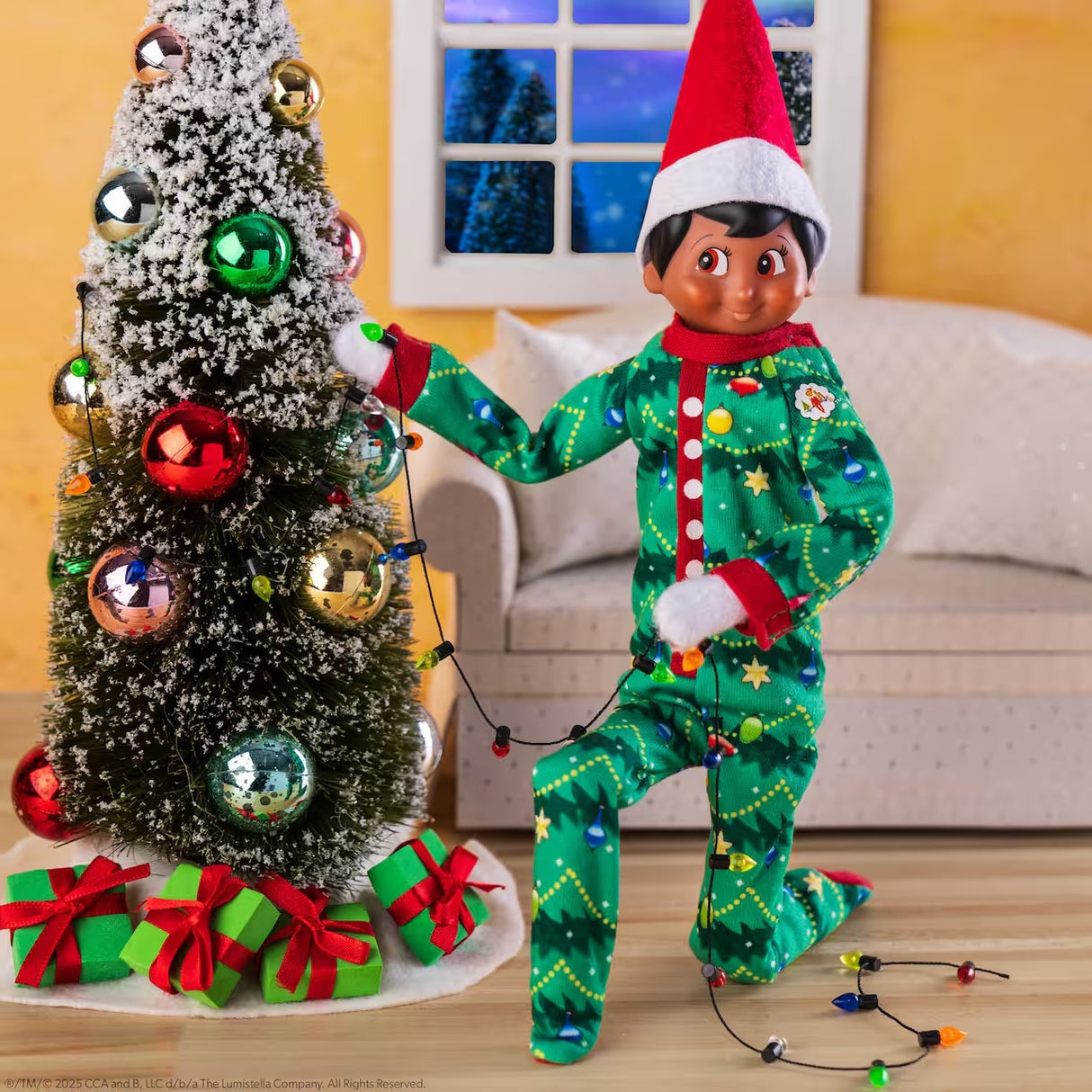 This is a lifestyle image showing a Scout Elf wearing the pajamas while decorating a small Christmas tree.