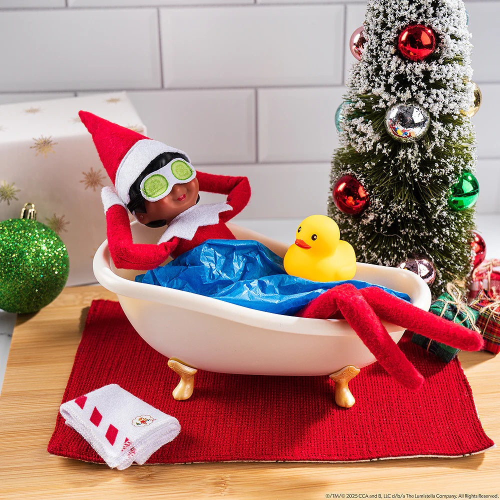 This is a lifestyle image showing a Scout Elf resting in a bathtub wearing the cucumber mask with the rubber ducky resting on him. The towel is on the floor next to the tub and there's a small tree next to it.