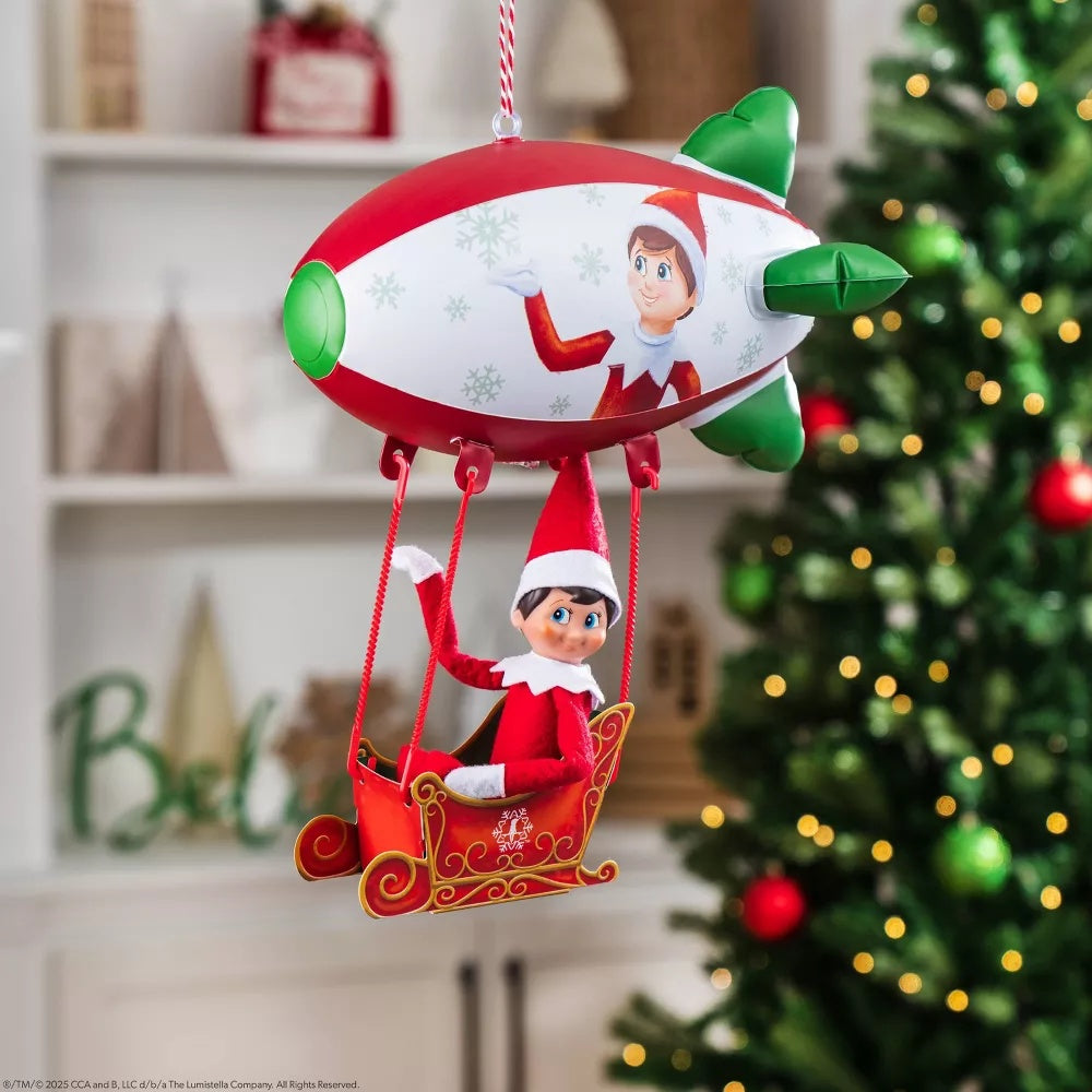 This is a lifestyle image showing the a scout elf sitting inside the sleigh underneath the blimp flying in front of a decorated Christmas tree.
