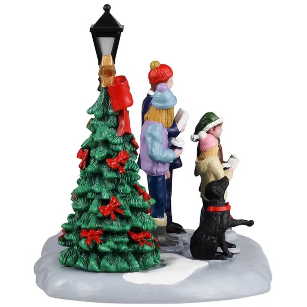 This shows a Lemax table accent from the side, designed to look like a family of four standing in snow in between a decorated Christmas tree and a lamp post. They're singing carols and their black dog howls with them.