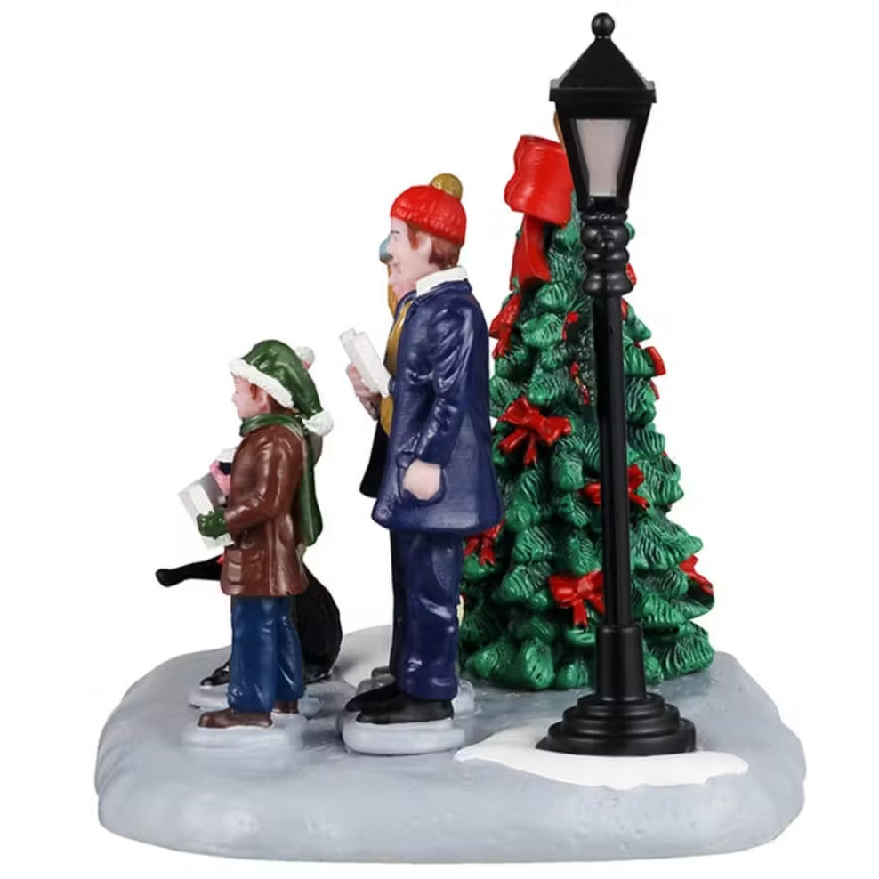 This shows a Lemax table accent from the side, designed to look like a family of four standing in snow in between a decorated Christmas tree and a lamp post. They're singing carols and their black dog howls with them.