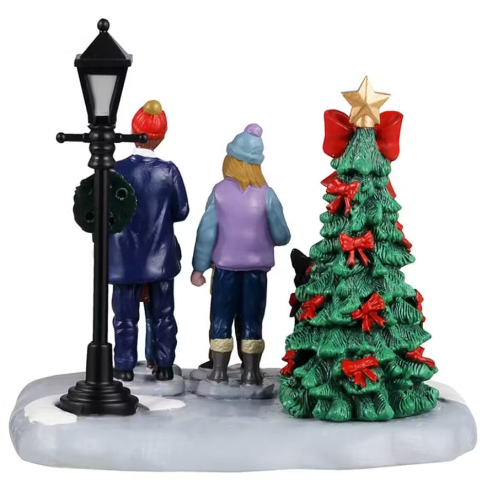 This shows a Lemax table accent from the back, designed to look like a family of four standing in snow in between a decorated Christmas tree and a lamp post. They're singing carols and their black dog howls with them.