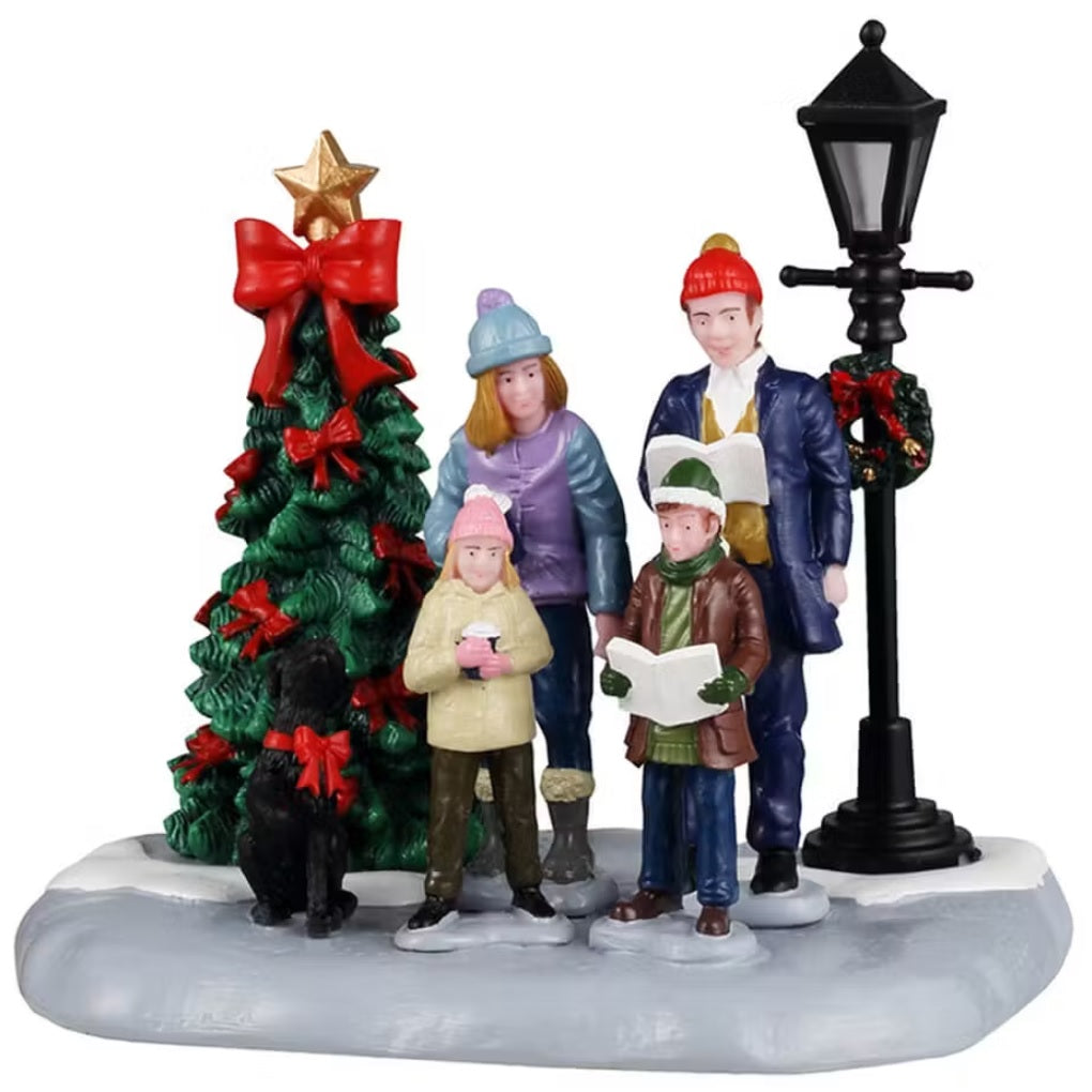 This shows a Lemax table accent designed to look like a family of four standing in snow in between a decorated Christmas tree and a lamp post. They're singing carols and their black dog howls with them.