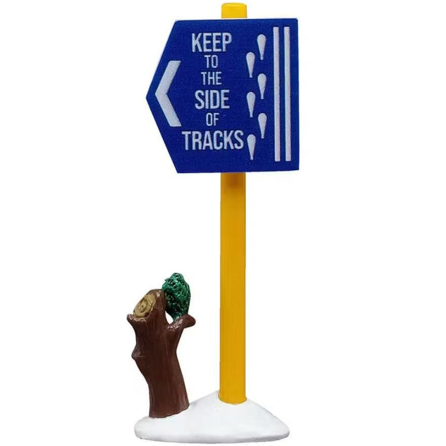 This is the sign that has a yellow pole and the blue sign reads "KEEP TO THE SIDE OF TRACKS" and has a small tree stump next to it.