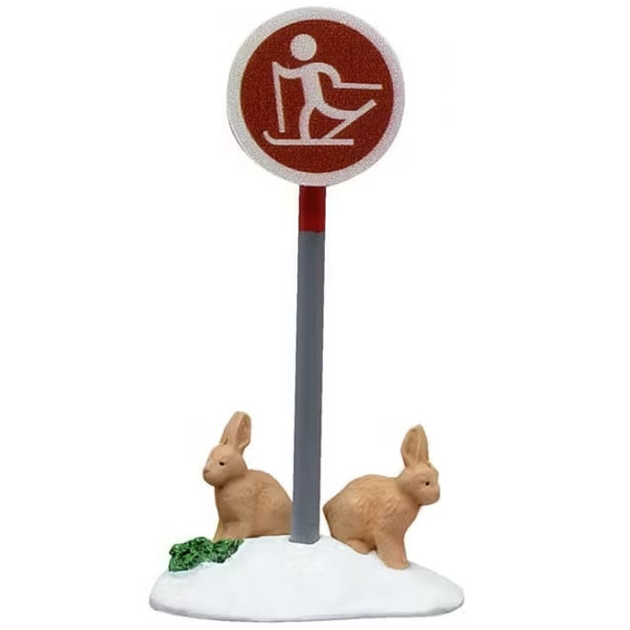 This is the sign that has a gray pole and a circular, red sign showing a cartoon person skiing and there are two tan colored bunnies underneath it.