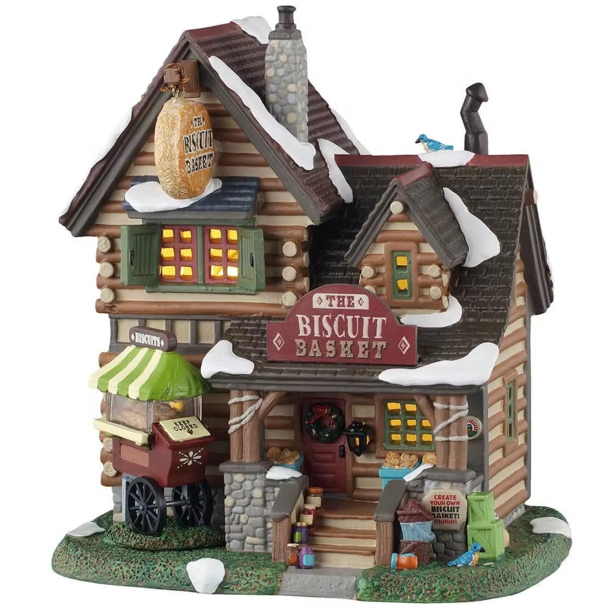 This is a Lemax lighted building designed to look like a tall log cabin. There's a blue bird on the roof and one on the grass near the front stairs. There's snow on the roof and on the grass. There's a sign by the stairs that reads "CREATE YOUR OWN BISCUIT BASKET!" There's a cart on the left of the stairs with biscuits inside. There are jars lining the stairs.