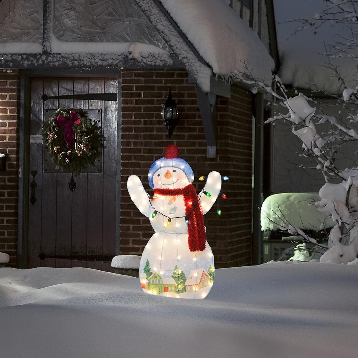 This is a lifestyle image showing the snowman sculpture on a snowy front lawn in front of a house decorated for Christmas.