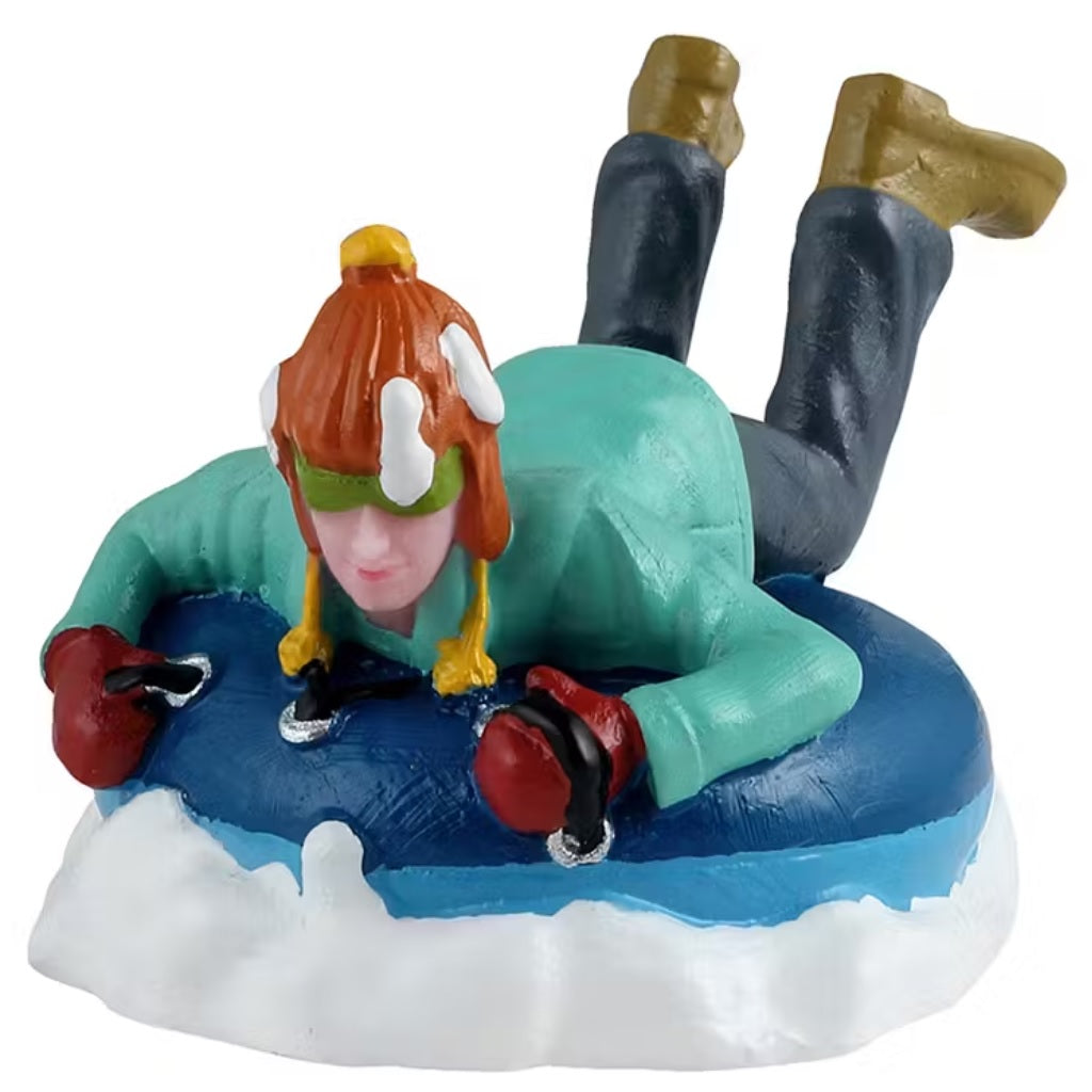 This is a Lemax figurine designed to look like a young boy laying on a circular, blue inner tube. He's wearing a teal jacket, dark blue pants and brown shoes. His orange hat and green goggles are covered in snow.