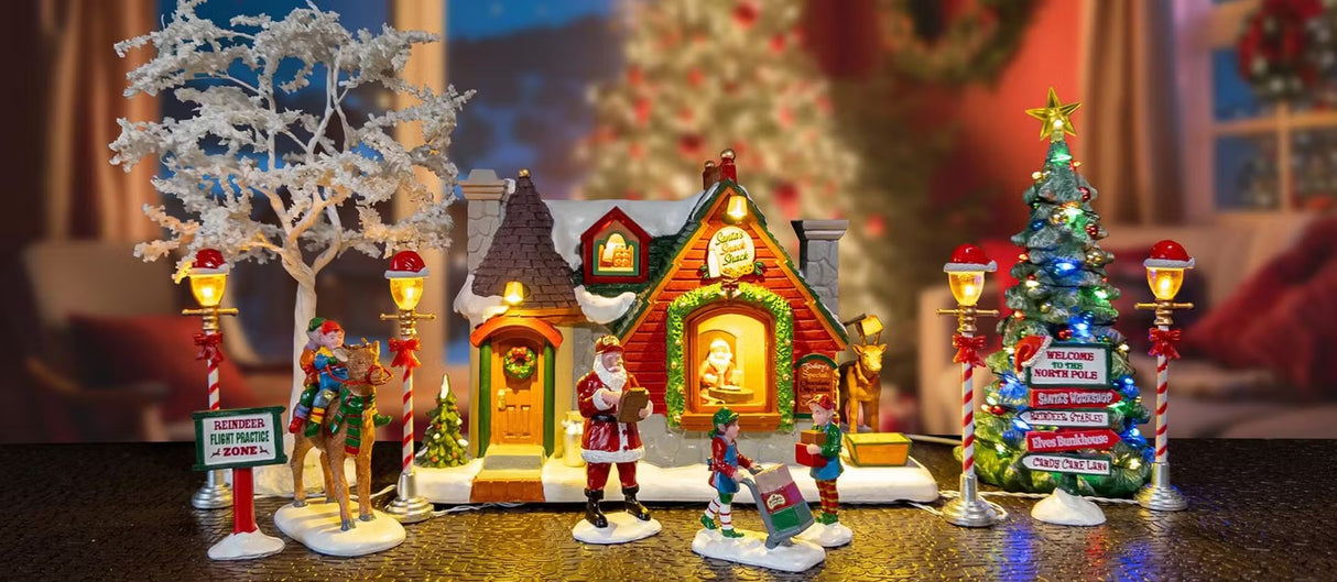 This is a lifestyle image showing this set on display. There's a tree on either side of the cottage and two lamp posts on either side, as well. There are two elves riding on a reindeer, Santa checking his list and two elves moving boxes. There are also village signs on either side of the display.