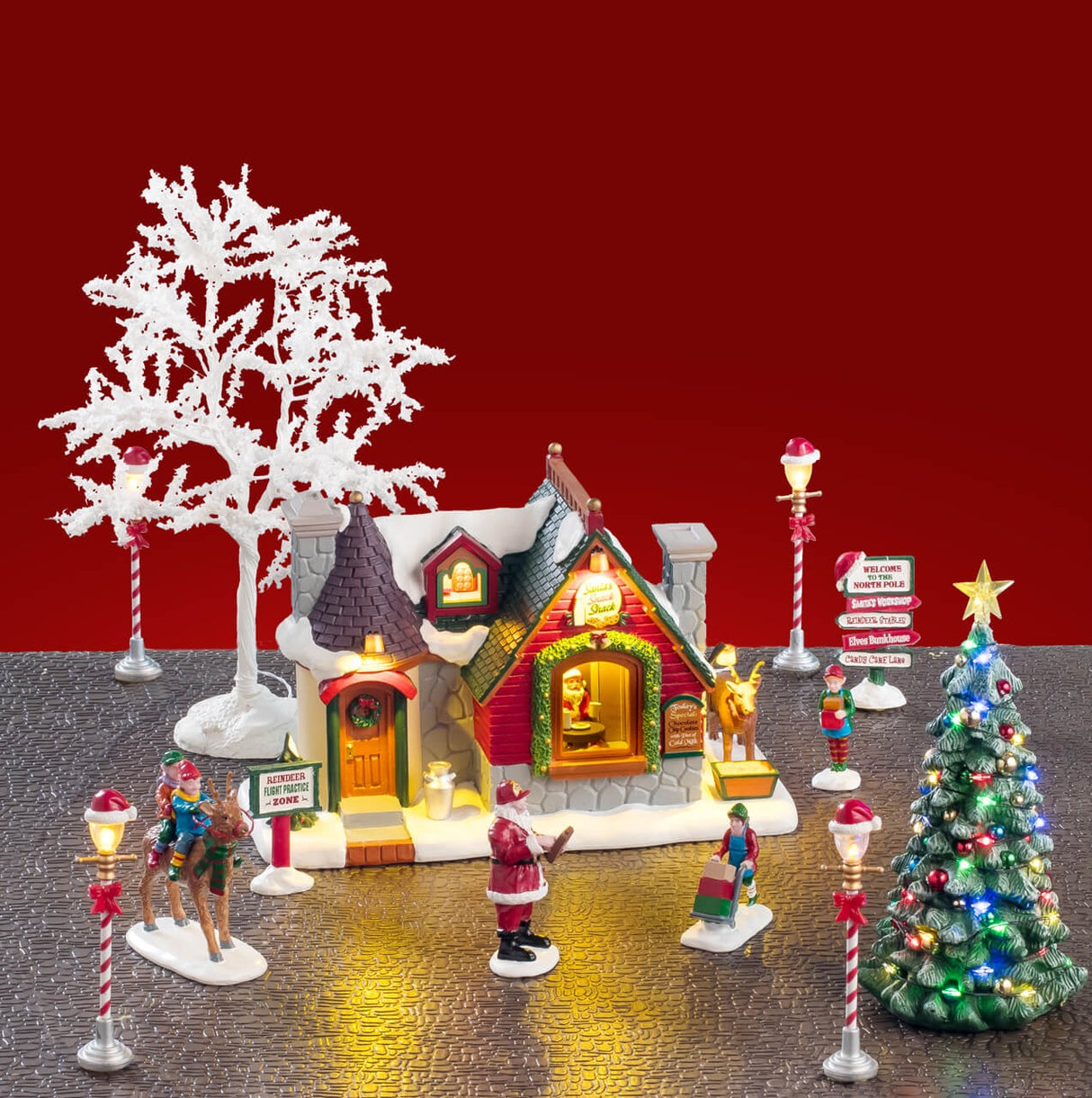 This is a lifestyle image showing this set on display. There's a white tree and two lamp posts behind the cottage.There are two elves riding on a reindeer, Santa checking his list and two elves moving boxes. There are also village signs on either side of the display and two lamp posts in front of the cottage.