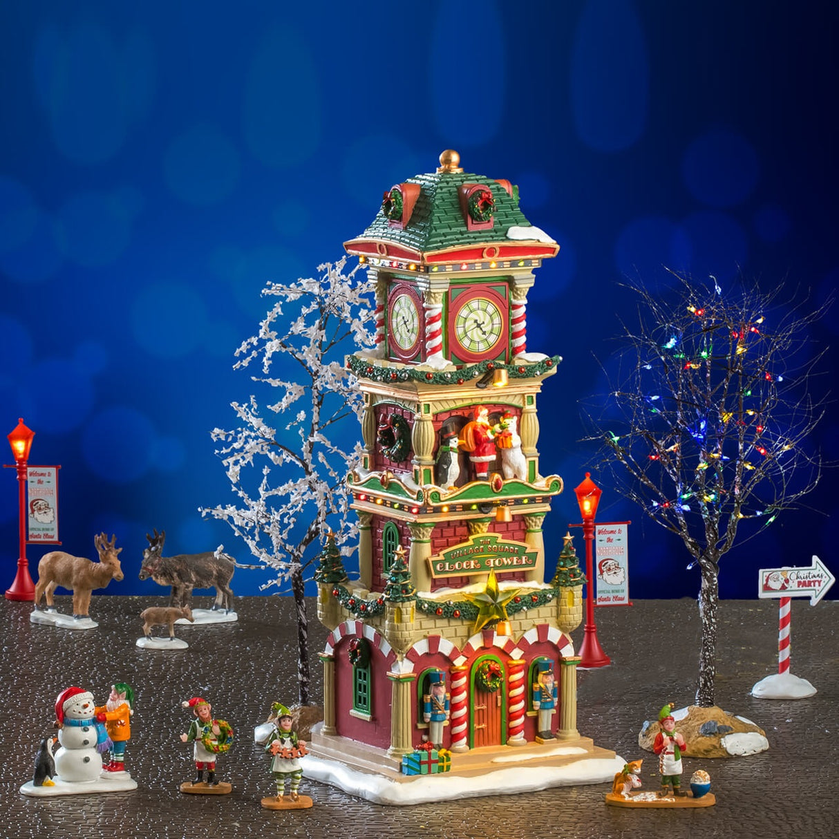 This is a lifestyle image showing this house set on display. There's the clock tower surrounded by two trees. The lamp posts are on either side of the building. In the back of the tower, there are three reindeer. In front of the tower, there's an elf and a penguin building a snowman and three elves holding homemade decorations.