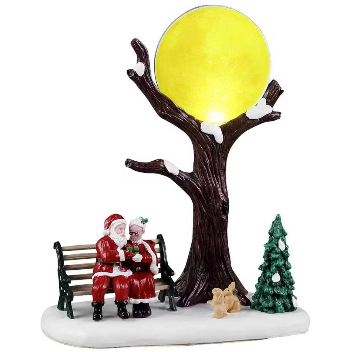 This is a Lemax table accent designed to look like Santa and Mrs. Claus are canoodling on a bench, sharing a present. The bench is next to a bare tree covered in snow, and the large moon is visible through the branches. There are two bunnies are staring up at the moon next to a tree.