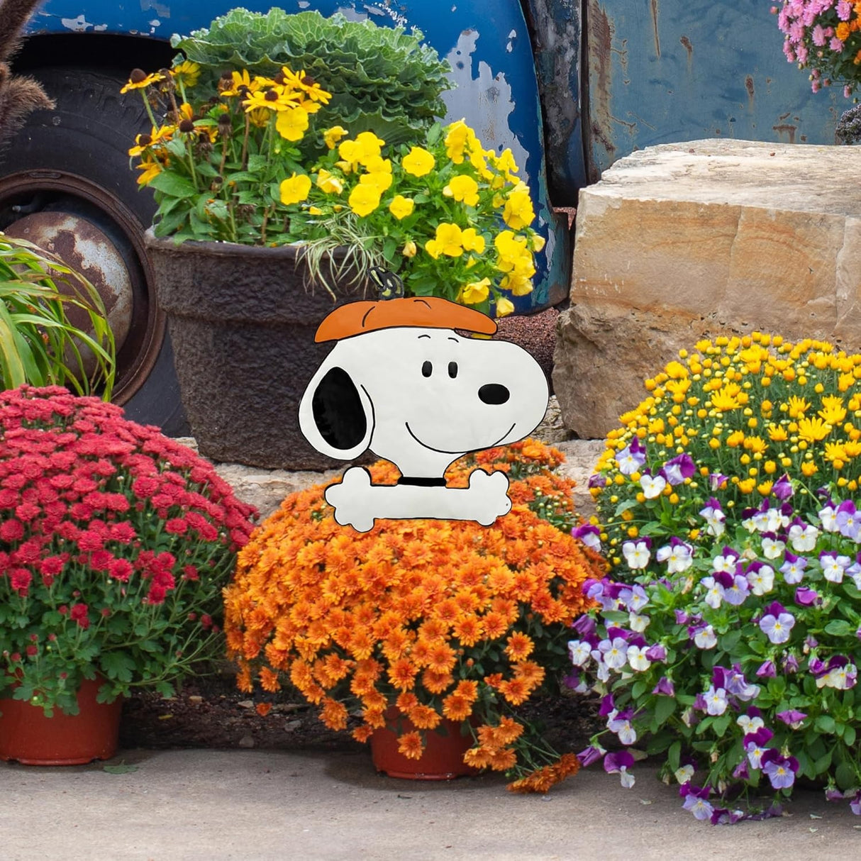 This is a lifestyle image showing off this pumpkin press-in decoration. It's sticking out of a pot of flowers. The pumpkin press-in decoration is designed to look like the famous black and white beagle from Peanuts cartoons: Snoopy. He's wearing the top of a pumpkin as a hat.