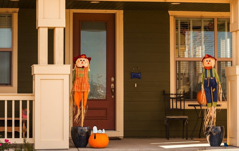 This is a lifestyle image showing the pumpkin press-in decoration in use. It's sticking out of a pumpkin on a porch. The porch is also decorated with two small scarecrows.