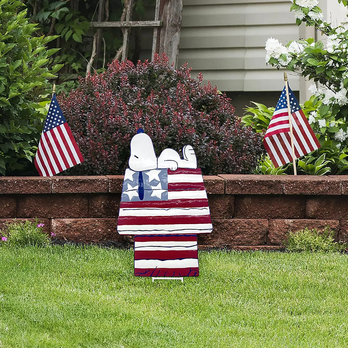 This is a lifestyle image showing this decorative garden stake in a garden. This decorative garden stake is designed to look like the famous Peanuts character, Snoopy. Snoopy is sleeping on top of his doghouse and the house is designed to look like the American flag: red, white and blue stars and stripes.