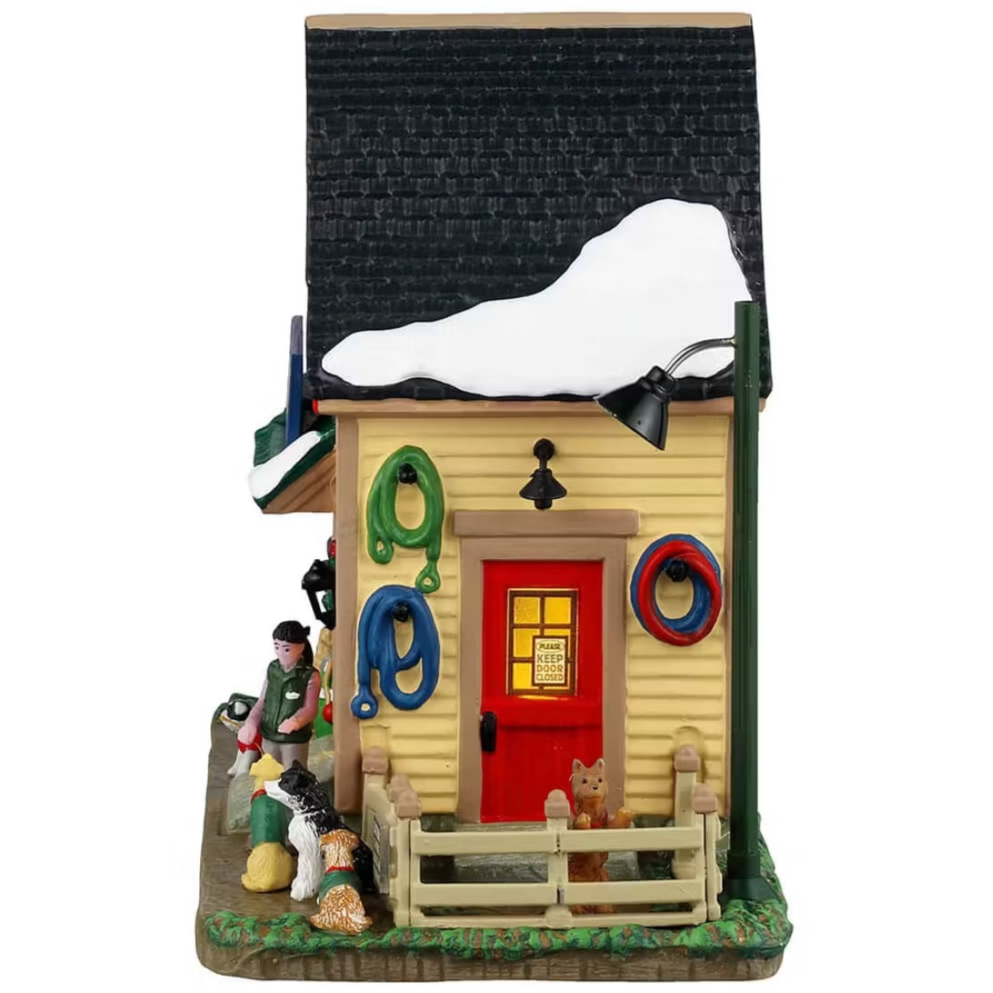 This is the side of the building. There's a red door and different colored leashes hanging on the side. There's a fence with a puppy inside. You can see a trainer with three different dogs.