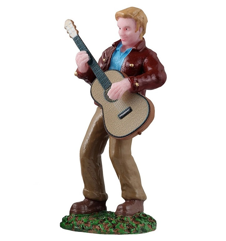 This is the figurine of a man playing guitar. He's got blonde hair, a maroon colored jacket, a blue shirt, brown pants and brown shoes. He's standing on grass.