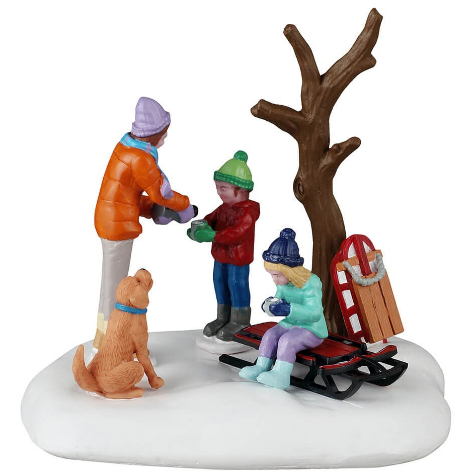 This is a Lemax table accent designed to look like a mom giving her kids hot chocolate while they rest from sledding next to a bare tree. There's a dog begging for some too.