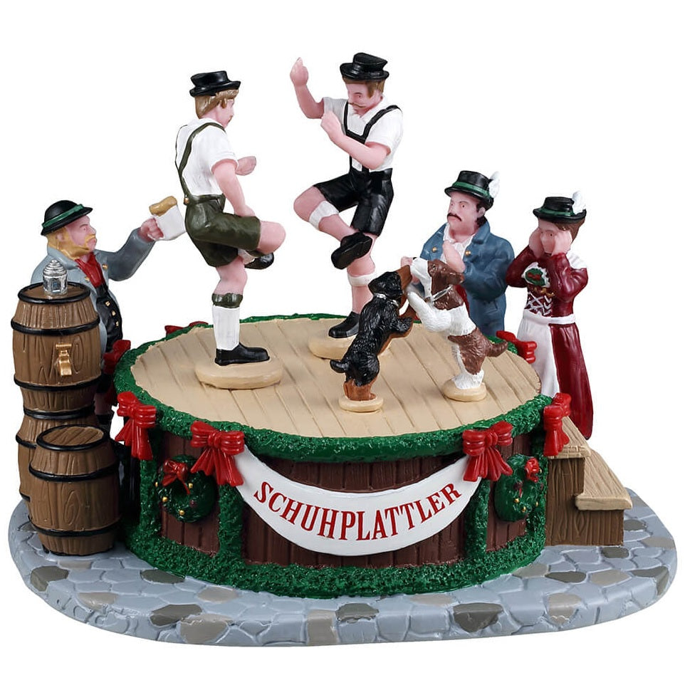 This is a Lemax table accent designed to look like men dancing on a wooden stage with two dogs. There's a banner on the side that reads "Schuhplattler." Schuhplattler is a traditional Bavarian and Tyrolean folk dance, known for its energetic movements and distinctive shoe slapping by the male dancers.