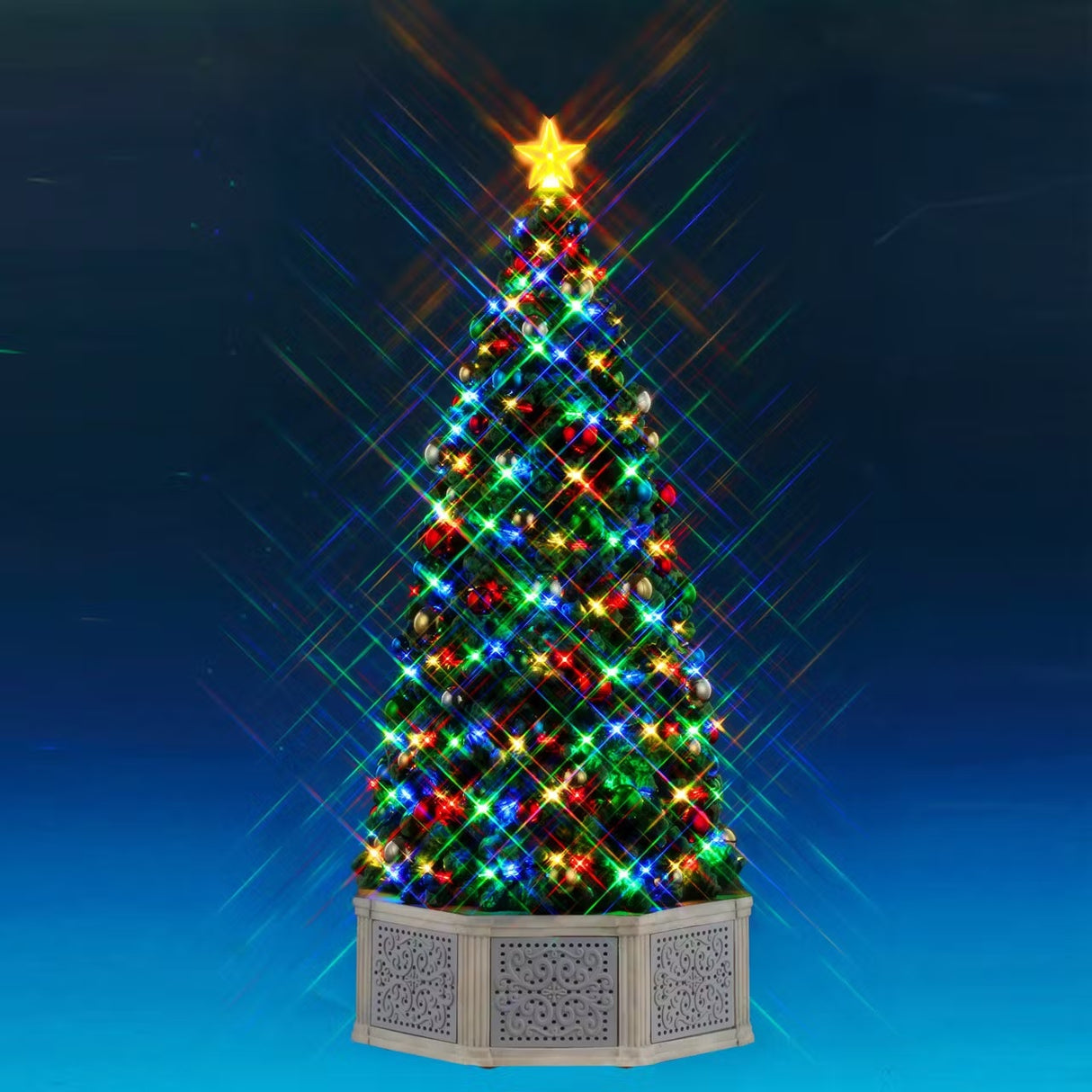 This is a Lemax lighted tree. There's a star on the top, lights throughout and shiny ornaments. There's a gray fence around the base of the tree. This shows the tree on a blue background.