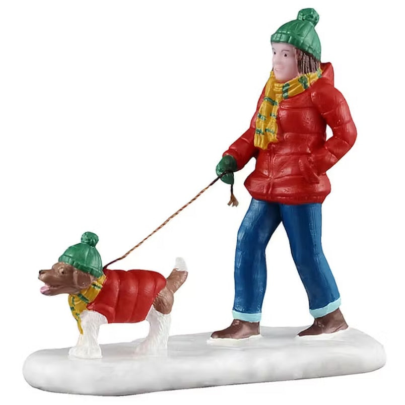 This is a Lemax figurine designed to look like a woman walking her dog on a leash in the snow. The woman is wearing a green hat, a red coat, a yellow and green scarf, green gloves, blue jeans and brown shoes. The little white and brown dog is matching her with a green hat, a yellow and green scarf and a red vest.