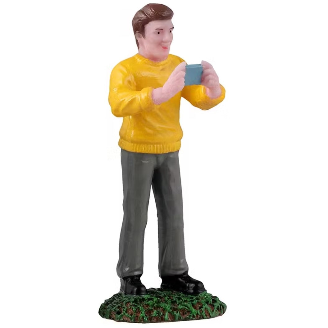 This figurine is a man wearing a yellow sweatshirt and gray pants and he's holding his phone as if he's recording.