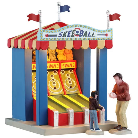 This is a Lemax table accent designed to look like a carnival skee ball booth. There are red and blue flags on the top and the roof was red and yellow. There are three skee ball stalls in the booth. A dad is giving his young boy one of the balls to throw into one of the skee ball holes. There are stuffed animal prizes on the top shelf of the booth.