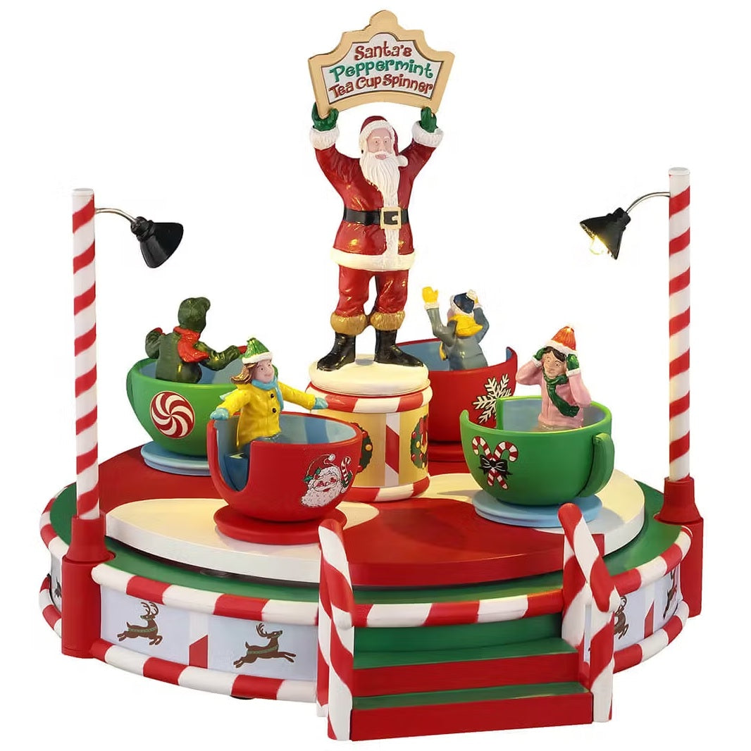 This is a Lemax table accent designed to look like a red and white striped carnival ride. In the center, there's a statue of Santa holding a sign that reads "SANTA'S PEPPERMINT TEACUP SPINNER." Around him, there are giant teacups with kids inside.