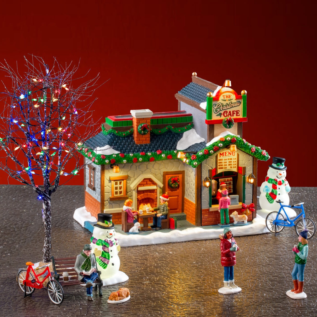 This is a lifestyle image showing this set on display. There's a cafe with a lighted tree on the left. In front of the tree, there's a bench with a man sitting on it. There's a bike leaning against the bench. There's a dog sleeping at the man's feet and a snowman next to him. There are two people having coffee. And on the right side of the cafe, there's a snowman and another bike.