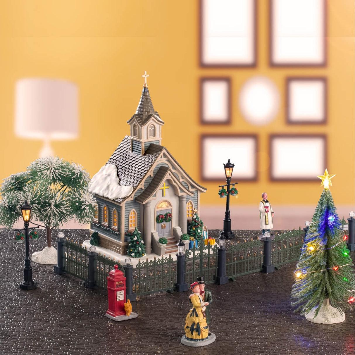 This is a lifestyle image showing this specific Lemax set. There's a church with a fence around it and a lamp post on both sides. There's a tree behind the church and a tree in front of the fence. There's a priest on the right side of the church, a red Victorian mail box with an orange cat at the base and a couple dressed in Victorian clothes strolling by.