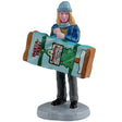 This is a Lemax figurine designed to look like a woman holding a box of an instant-tree. The box reads "INSTA-TREE! Everlasting Christmas Trees!" She's wearing a light blue winter hat and scarf, a blue jacket, a yellow shirt and gray pants.