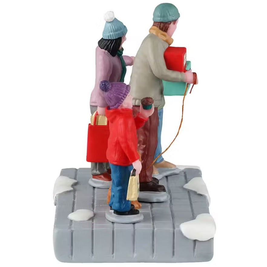 This is the side of a Lemax table accent that is designed to look like a family of four wearing coats, winter hats and scarves standing on a snowy sidewalk. They're all holding boxes. The young son is holding a box that reads "FREE SAMPLES." The dad is holding two boxes: "Dog Treats" and "Christmas Cake Factory." The mom is holding a red bag. The young daughter is holding a box that reads "Christmas Cake Factory." The dad is also holding the leash of their dog who is wearing a Santa hat.