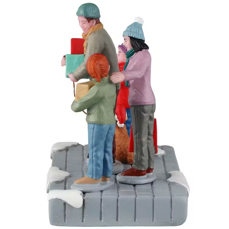 This is the side of a Lemax table accent that is designed to look like a family of four wearing coats, winter hats and scarves standing on a snowy sidewalk. They're all holding boxes. The young son is holding a box that reads "FREE SAMPLES." The dad is holding two boxes: "Dog Treats" and "Christmas Cake Factory." The mom is holding a red bag. The young daughter is holding a box that reads "Christmas Cake Factory." The dad is also holding the leash of their dog who is wearing a Santa hat.