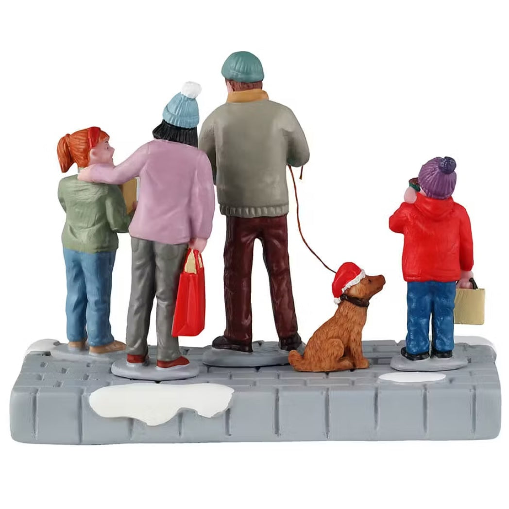 This is the back of a Lemax table accent that is designed to look like a family of four wearing coats, winter hats and scarves standing on a snowy sidewalk. They're all holding boxes. The young son is holding a box that reads "FREE SAMPLES." The dad is holding two boxes: "Dog Treats" and "Christmas Cake Factory." The mom is holding a red bag. The young daughter is holding a box that reads "Christmas Cake Factory." The dad is also holding the leash of their dog who is wearing a Santa hat.