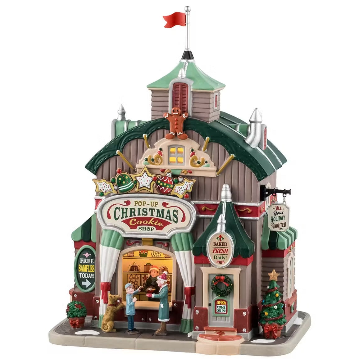 This is a Lemax lighted building called the Pop Up Christmas Cookie Shop. Two people at the front are enjoying cookies while a dog tries to steal one from the counter. There are Christmas decorations and wreaths and decorated trees.