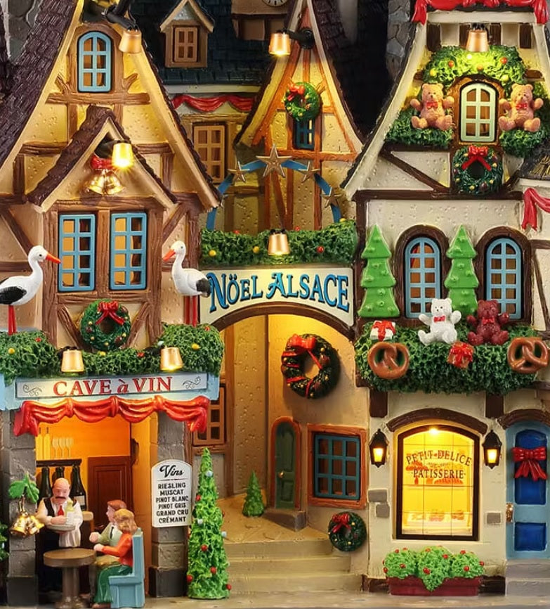 This is an up close view of the building. There's a restaurant on the left; a waiter is taking a table's order. Above one of the other shops, there's a garland decorated with gifts, pretzels and teddy bears.
