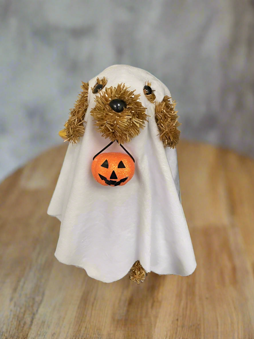 This tabletop decoration is a brown tinsel dog with black eyes and a black nose. He's wearing a soft, white sheet with eyeholes and a hole cut for his snout - so he looks like a ghost! He's holding a pail in his mouth that's shaped like a jack-o-lantern.