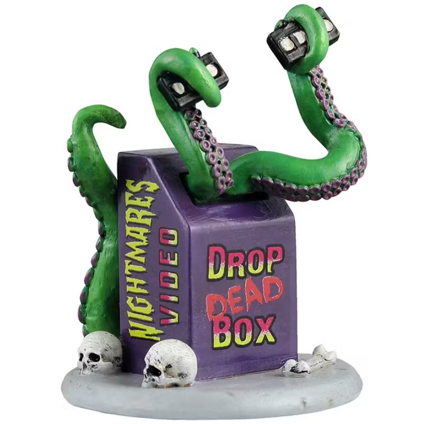 This is a Lemax Spooky Town accessory designed to look like a purple video return box. On one side it reads "NIGHTMARES VIDEO" and on the other side it reads "DROP DEAD BOX." There are green monster tentacles coming out of the box holding VHS tapes. There are skulls and bones at the base of the box.